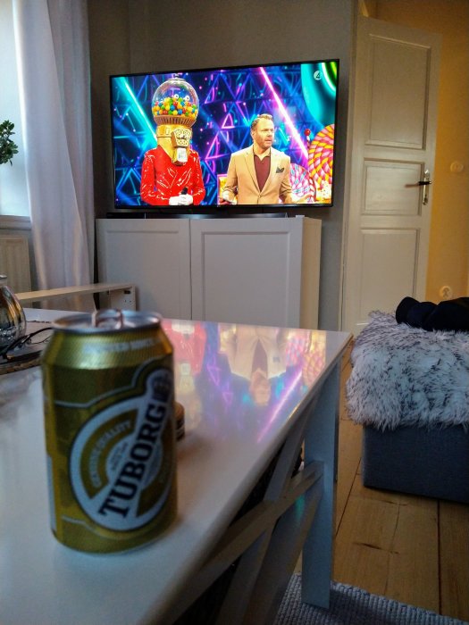 En Tuborg Gold-ölburk i förgrunden med en TV som visar Masked Singer i bakgrunden.