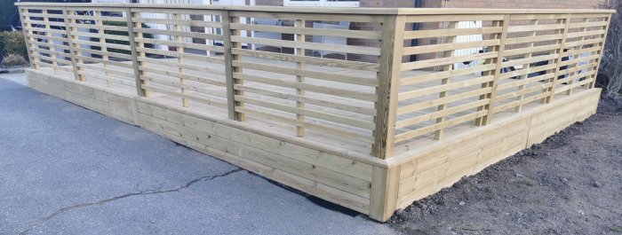 Nybyggd träterrass med integrerat staket av träribbor längs en betongväg.
