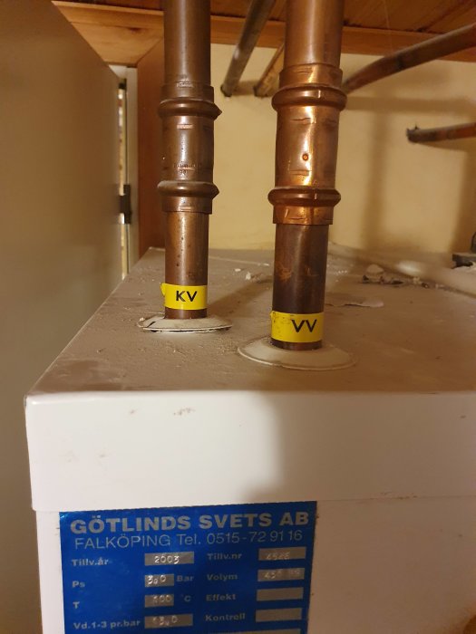 Kopparledningar märkta med 'KV' för kallvatten och 'VV' för varmvatten på en vit vattenbehållare.