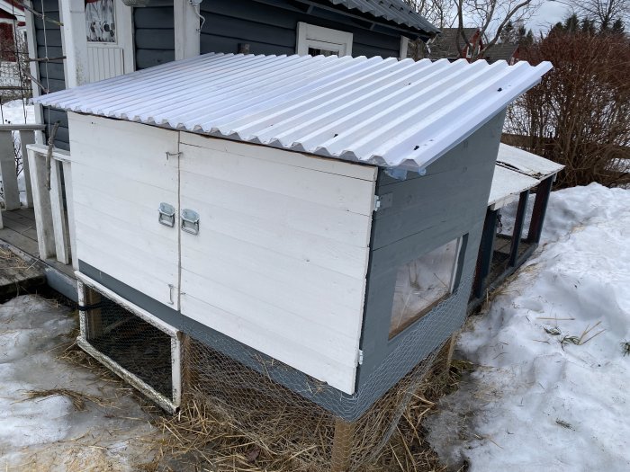 Ett hemmabyggt isolerat djurhus med vit sidopanel, vågigt tak och en glaslucka från en TV-bänk för ljusinsläpp, beläget på ett snöigt område.