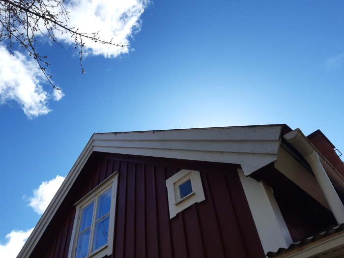 Takdetalj av ett rött timmerhus med vitt snedtak, tegelpannor och hängrännor mot en klarblå himmel.