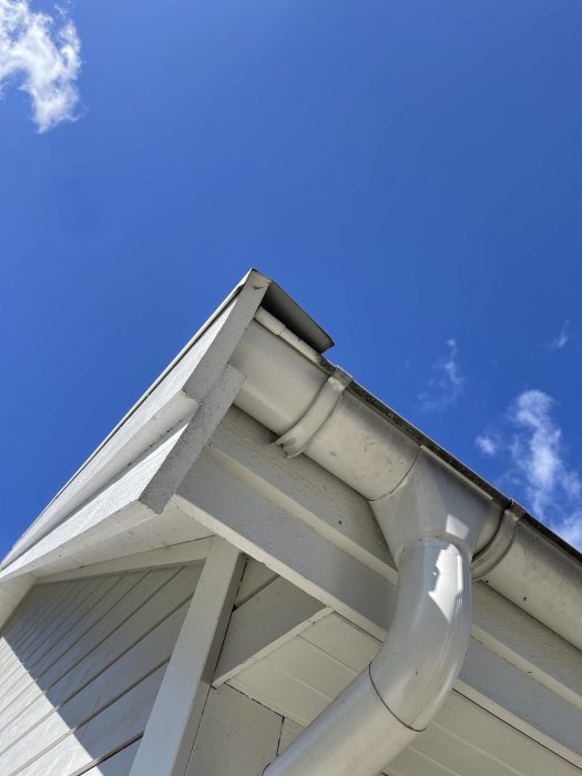 Hörn av hus med vattplåt som sticker ut utanför hängrännan mot en klarblå himmel.