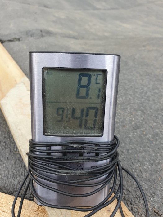 Digital termometer som visar 8.1°C utomhus och 95.4% luftfuktighet, placerad på träyta med kabel.