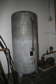 En gammal varmvattenberedare i metall med mätare och rörledningar i ett övergivet utrymme.