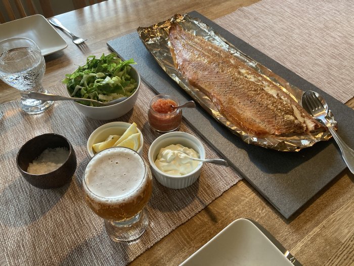 Helstekt fisk på folie, serveringstillbehör såsom citron, sås och sallad, samt ett glas öl på matbord.