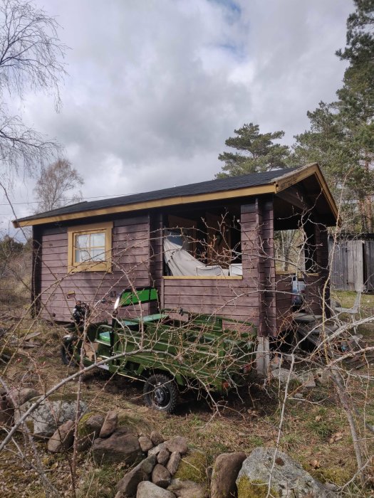 En grön trädgårdstraktor parkerad vid en brun trästuga med schingeltak och synliga fönster.