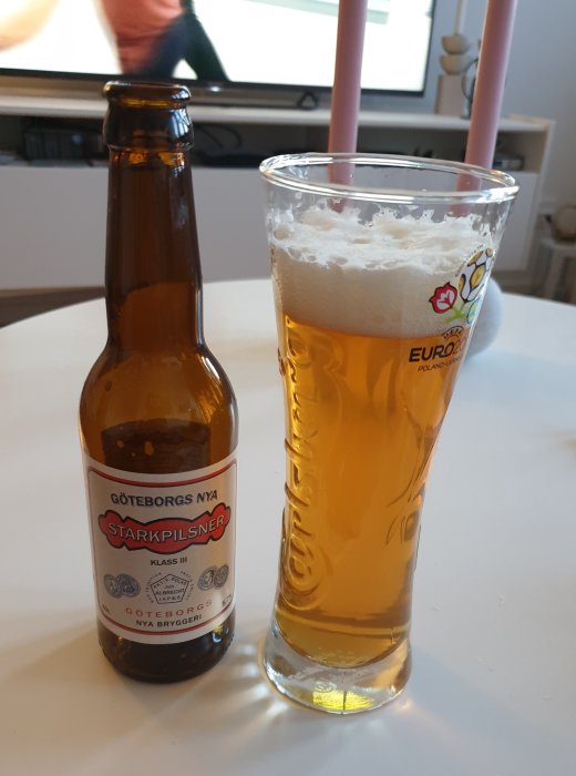 En öppnad ölflaska och ett glas med öl från Göteborgs Nya Bryggeri på ett bord framför en TV.