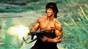 En vältränad man som avfyrar ett maskingevär, filmisk heroisk pose, solbränd hud och bar överkropp.