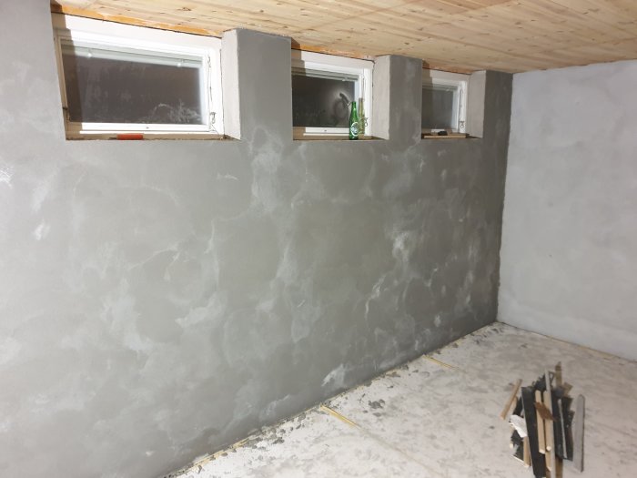 Vägg under putsning med exponerade fönstersmygar och en hög med brädor på golvet.