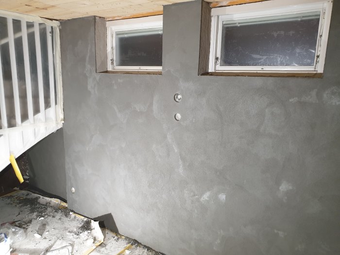 Vägg under putsarbete med delvis putsade fönstersmygar och byggavfall på golvet.
