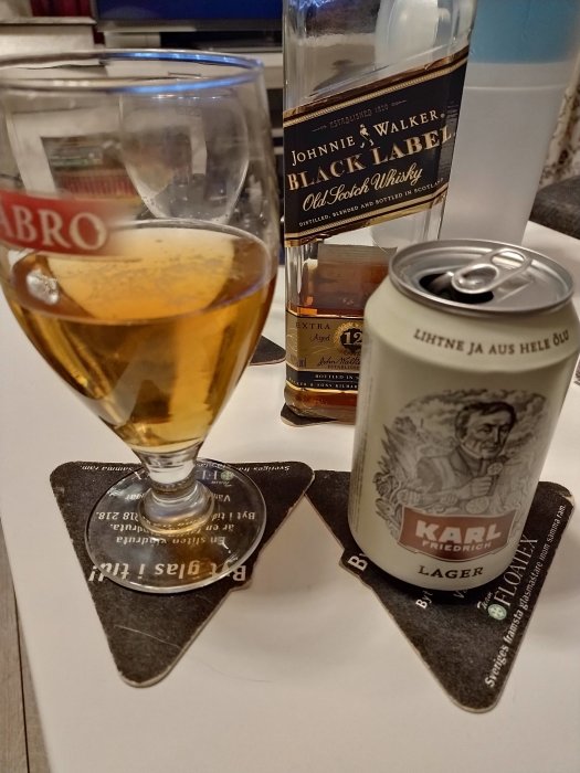 Ölglas med dryck på bordsunderlägg samt flaskor av Johnnie Walker whisky och burk av Karl Friedrich lager.