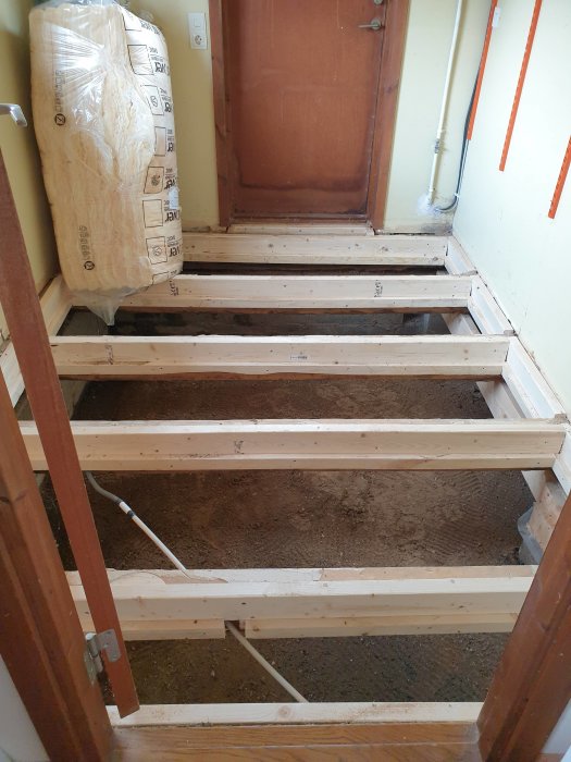 Nybyggda träreglar på ett golv under renovering, med isolering i förpackning och rör längs väggen.