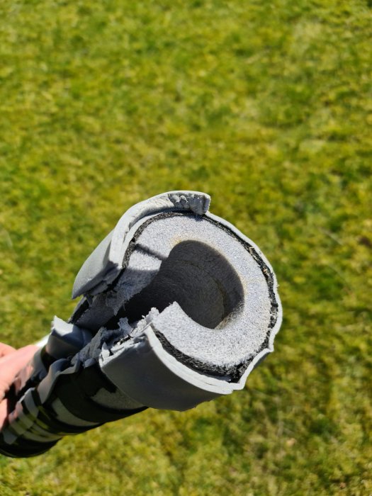 Isoleringsmaterial klippt till för att isolera en rörböj, hållen mot en grön gräsmatta i bakgrunden.