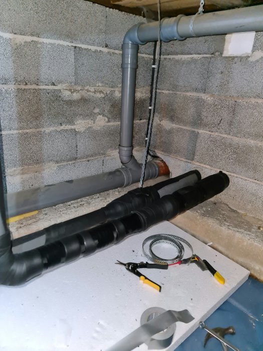 Isolerade rör i en krypgrund med verktyg och isoleringstejp synlig på vit isolerskiva.