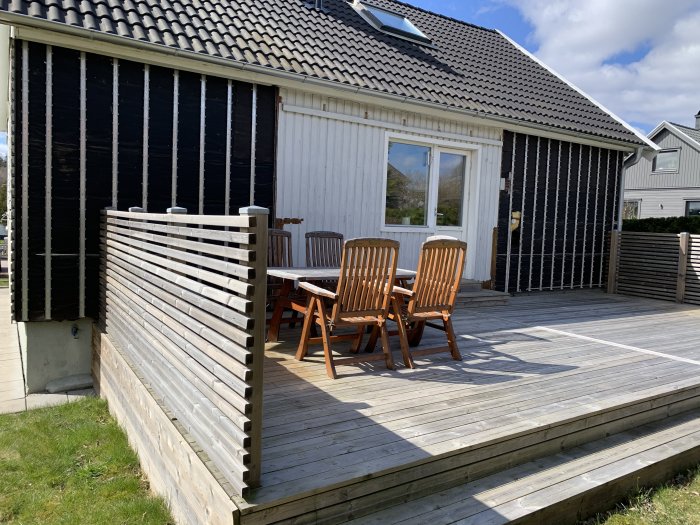 Hus med delvis borttagen fasadsten och träpanel, träterrass med bord och stolar under soligt väder.