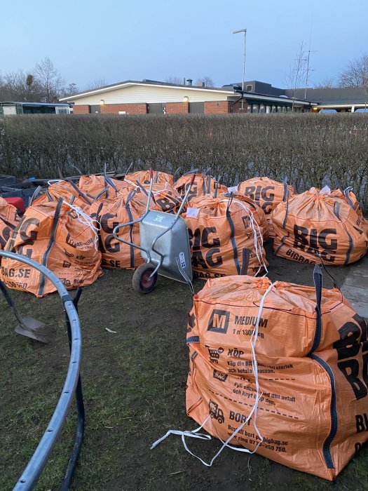 Flera orangea bigbags fyllda med jord på en trädgård med en skottkärra och verktyg i förgrunden.