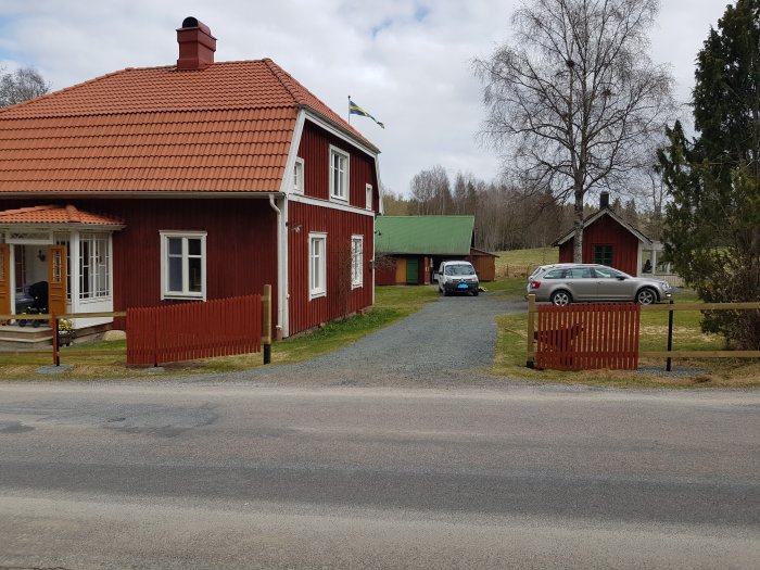 Rödmålat trähus med torkställning framför, grusuppfart och slänt med ribbor och två bilar.