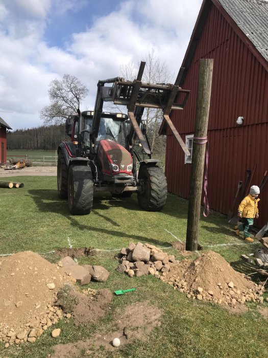 Traktor lyfter ner stolpar i grävda hål för nytt plank, med utgrävda jordhögar och sten omkring.