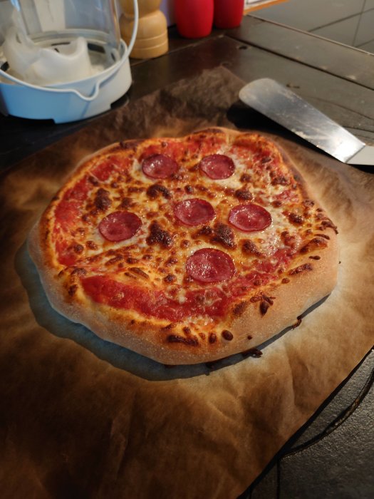 Nybakad pizza med salami på bakplåtspapper och en degkrok i bakgrunden.