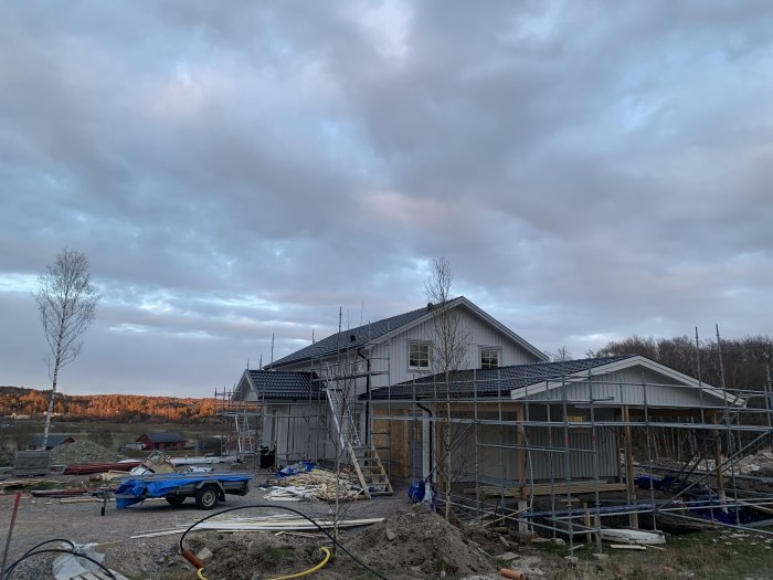 Hus under konstruktion med byggnadsställningar och material på marken under en molnig himmel.