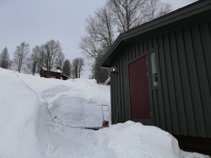 Vinterstuga med tjocka snödrivor runtom, upplyst trädgårdslampa och en röd dörr.