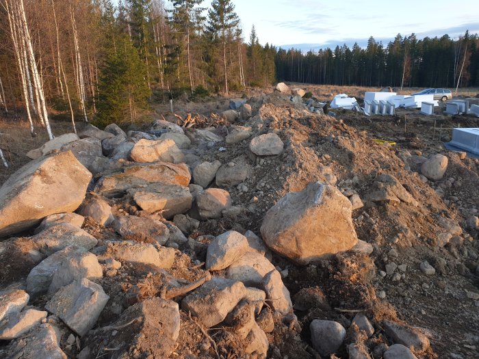 Stor hög av schaktmassor bestående av morän och stenar vid skogsbryn, nära byggarbetsplats.