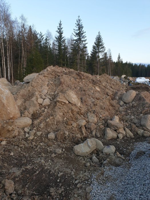 Hög med fyllnadsmassor och större stenar från grundarbete, med skog i bakgrunden.