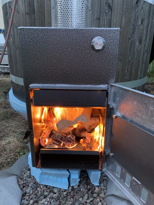 Eld brinner i en vedeldad badtunnas värmepanna med öppen lucka, intill en träbeklädd tunna.