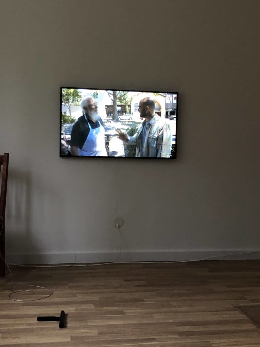 TV monterad på vägg ovanför ledningsrör och lösa kablar på golvet, två personer visas på skärmen.