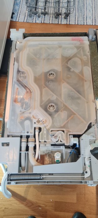 Öppen diskmaskin på golvet som visar insidan med demonterad plasttank, avloppsslang och synliga vattenkanaler.