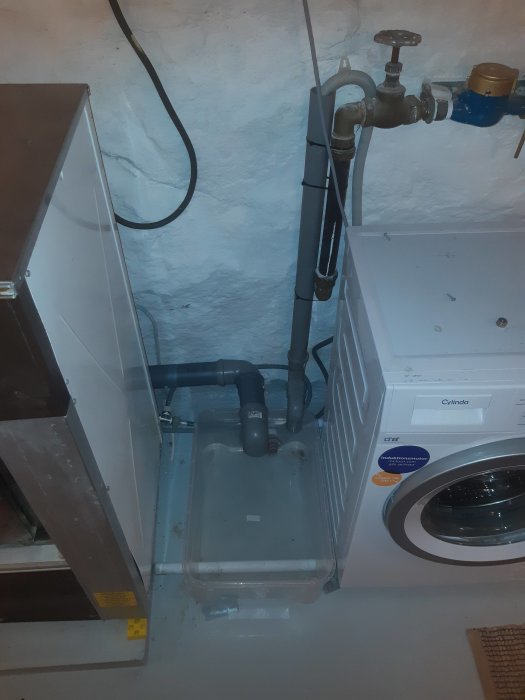 Vatteninstallation i källare utan filter, inkluderar rörledningar, tvättmaskin och transparent behållare.