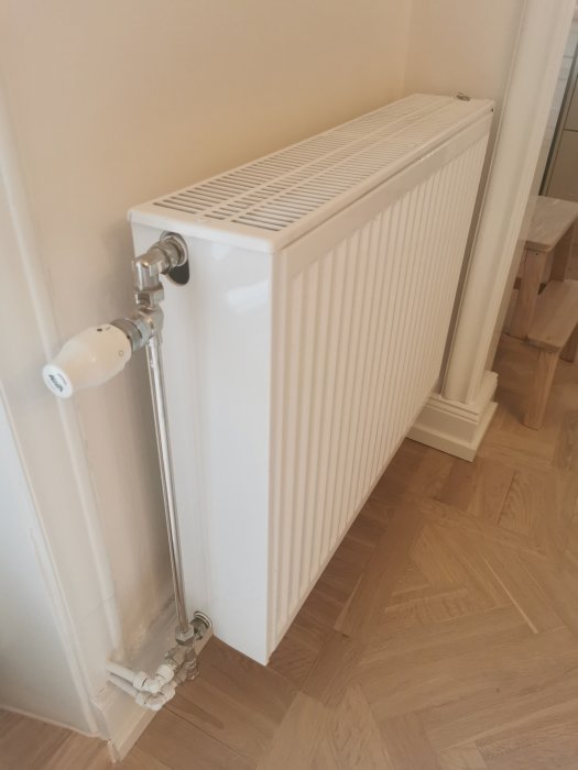 Vit modern radiator med termostatventil monterad vid vägg i ett hörn på ett trägolv.