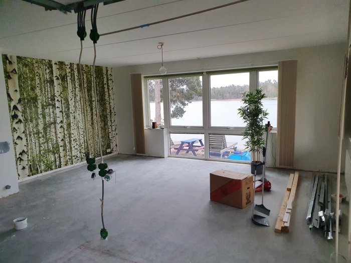 Tomt renoveringsutrymme med betonggolv, plåtreglar, ledningar och utsikt över sjö.