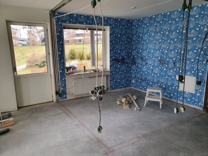 Tomt rum under renovering med blått tapetserat vägg, betonggolv och elektriska ledningar synliga.