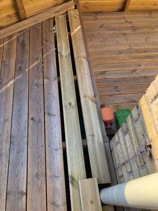 Nybyggd trätrappa med obehandlade steg på en veranda, nära en tegelvägg och vit stuprör.