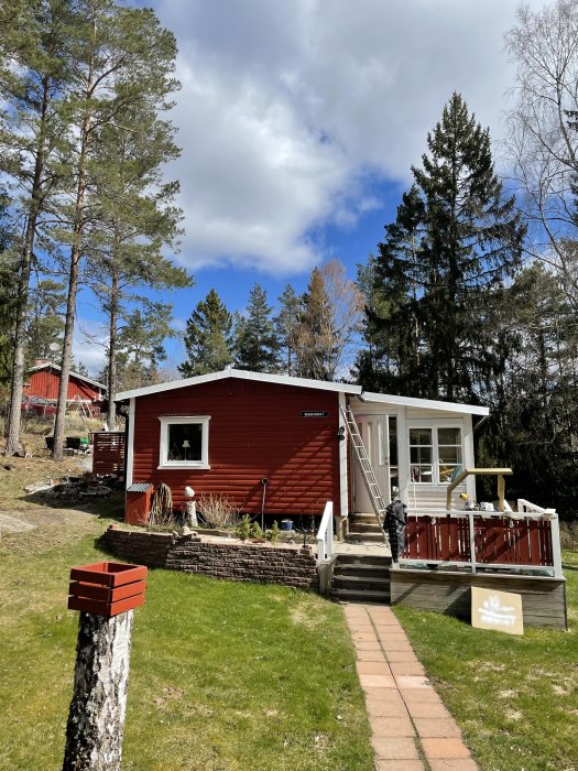 Rött fritidshus med vita fönster och veranda i en trädgård omgiven av träd under blå himmel.