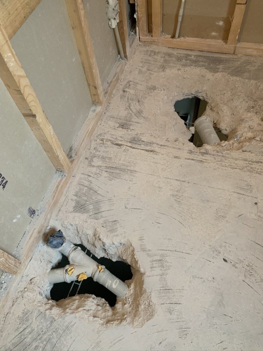 Puuteräkenteinen lattia, jossa kaksi reikää ja näkyviä muoviputkia, kertoo rakentamisen tyylistä ja keskeneräisestä remontista.