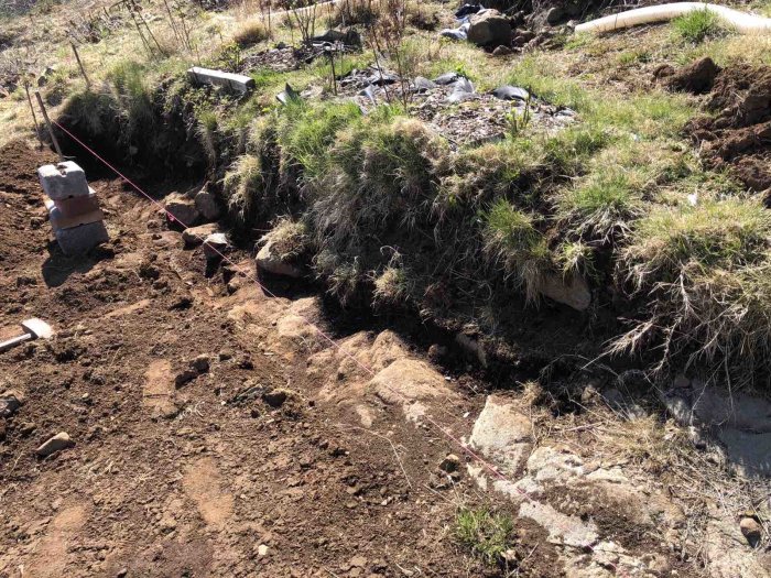 Område med utgrävd slänt och utspridda stenar inför byggandet av en stödmur.