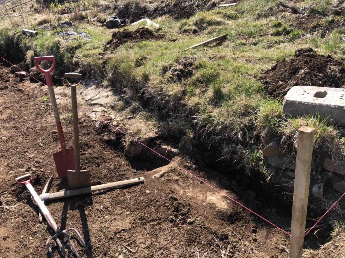 Påbörjad grävning för stödmur med redskap, ojämn terräng, berg och marklinjer synliga.