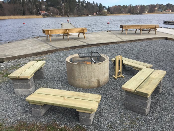 Nybyggd eldplats med bänkar av trä och betong vid en sjö med brygga i bakgrunden.