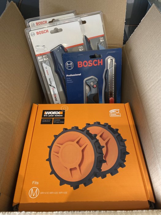 Öppen kartong innehållande nya verktyg; BOSCH sågblad och knivar samt ett paket med orangea off road-hjul från WORX.