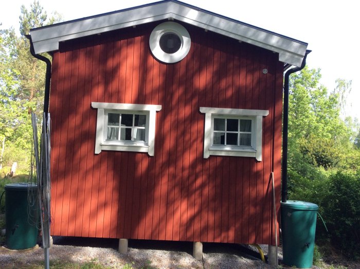 Rött trähus med vitt runt fönster högst upp och två kvadratiska fönster på var sida, stående i grönska.