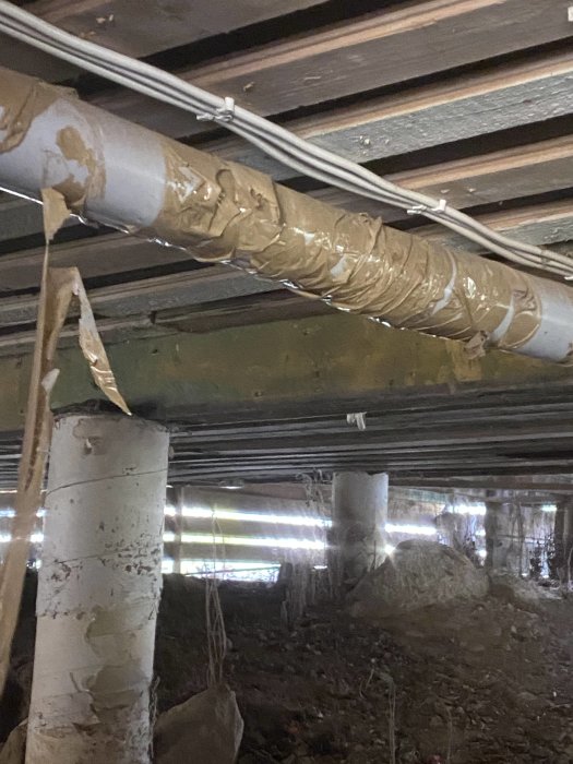 Undergolv med synliga bjälkar, isolerade rör och pelare i ett potentiellt torpargrund.