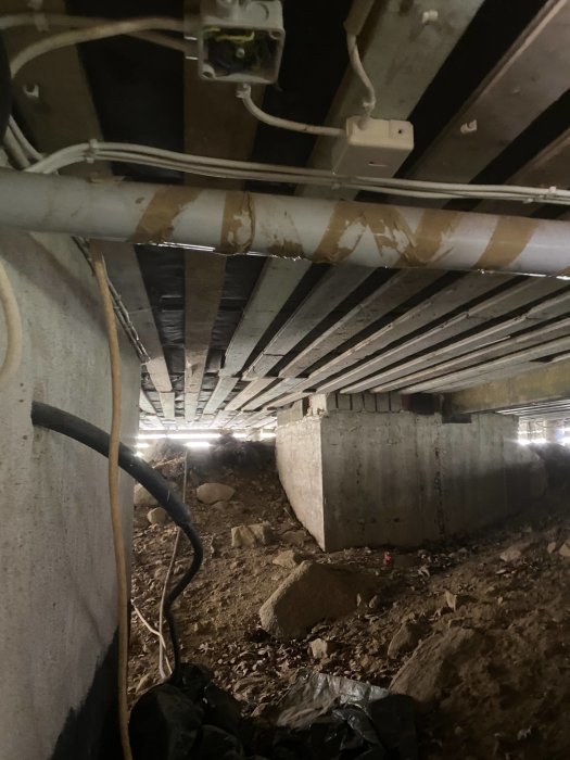 Vy under ett torp med torpargrund som visar träbjälkar, stödmurar i betong och installationer av rör och kablar.
