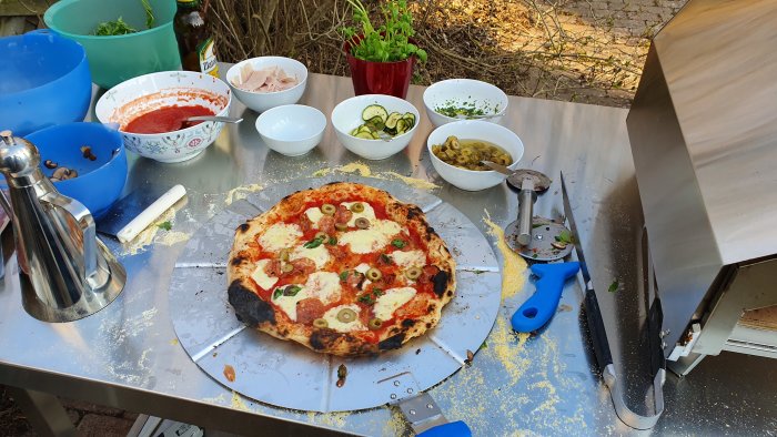 Hembakad pizza med olika toppings intill pizzaugnen och ingredienser utomhus.