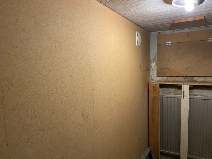 Källarrum med brun väggskiva, ett äldre vedinskast till höger och en del av en radiator, i behov av renovering.