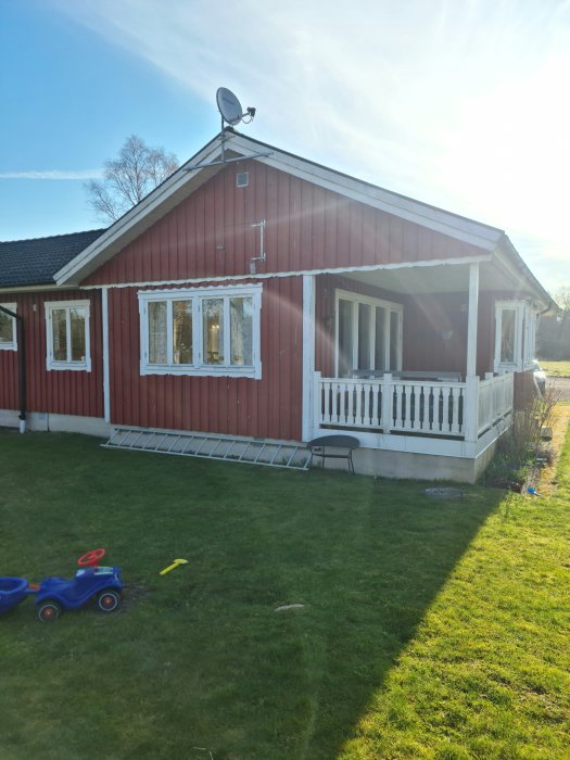 Rödmålat trähus med vita fönster och veranda, satellitantenn på taket, grön gräsmatta i förgrunden.