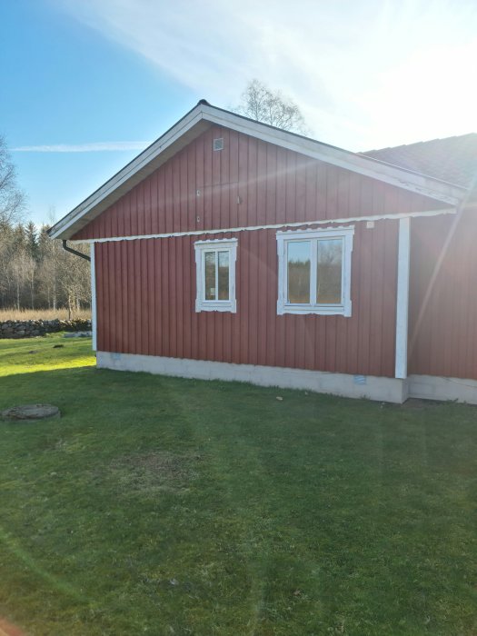 Rödmålad träfasad på ena sidan av ett hus med två vita fönster och vita knutar under solig himmel.