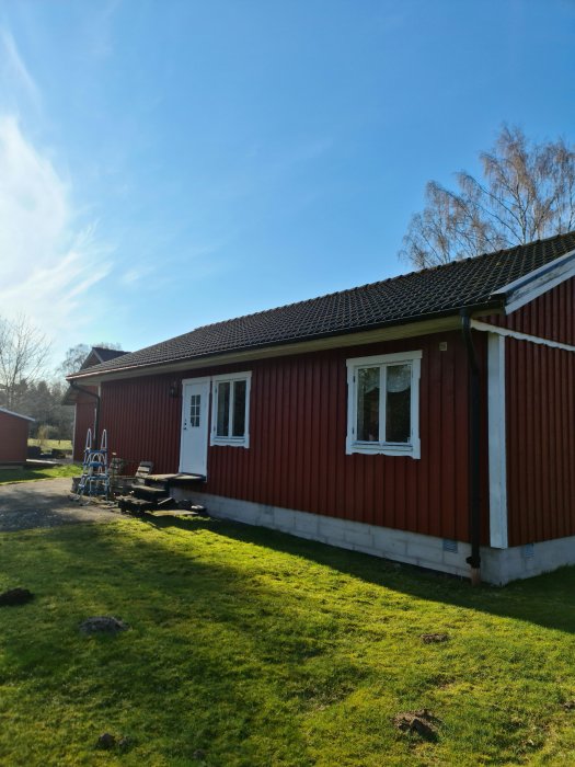 Rött trähus med vit knut och fönsterfoder, grönt gräs i förgrunden och solig himmel ovanför.