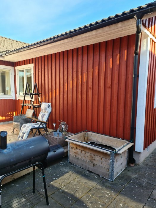 Rödmålat hus med träfasad och vita fönsterkarmar, en stege lutad mot väggen och en grill framför huset.